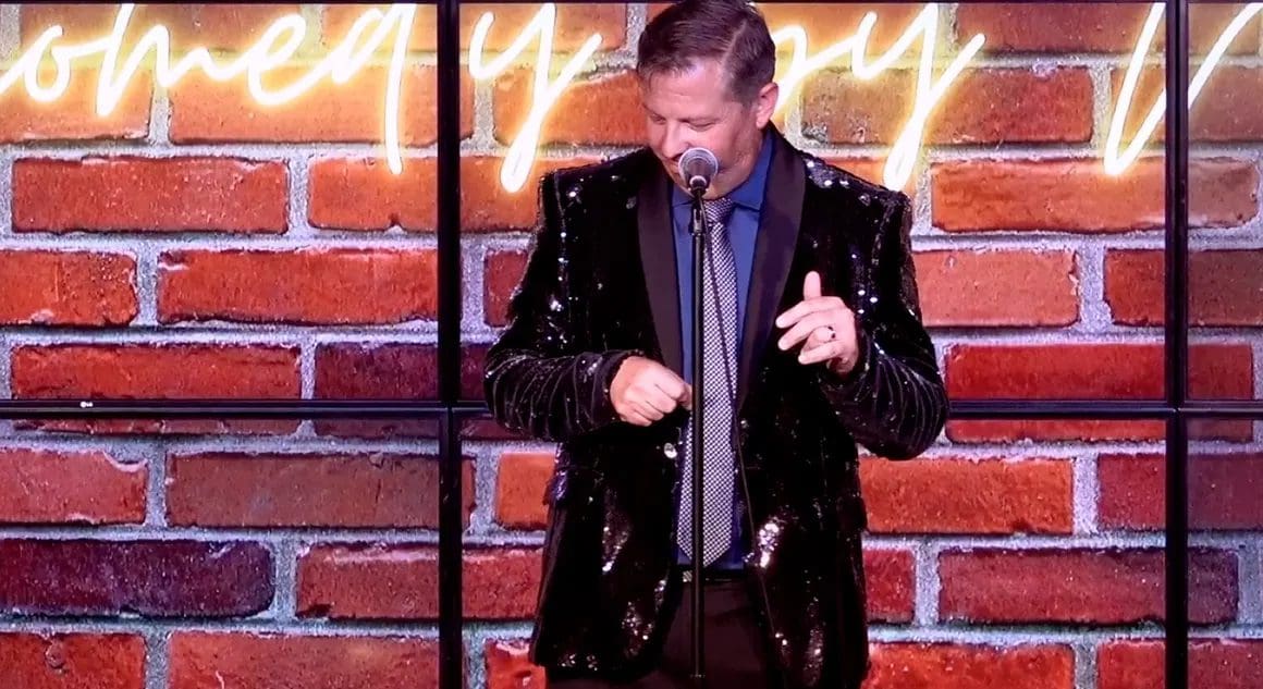 A man in black jacket and tie standing on stage.