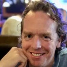 A man with curly hair and a blue shirt is smiling.